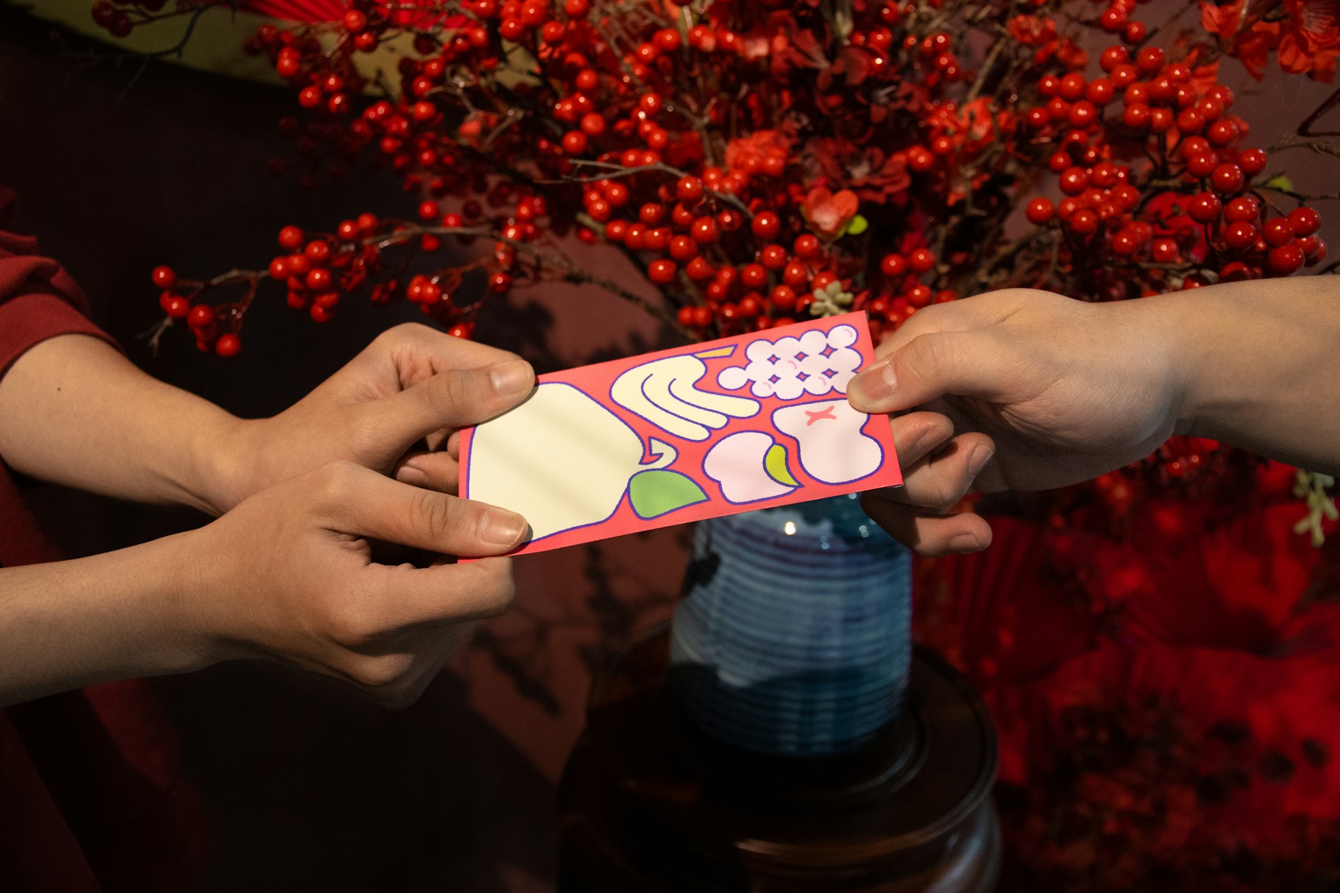 Hands exchanging a traditional Vietnamese lì xì red envelope with decorative designs against festive red berry background during Tết celebration