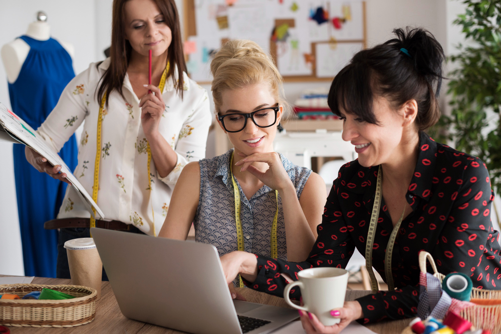Three women collaborating in a creative workspace with colorful design materials, dress forms, and casual patterned clothing